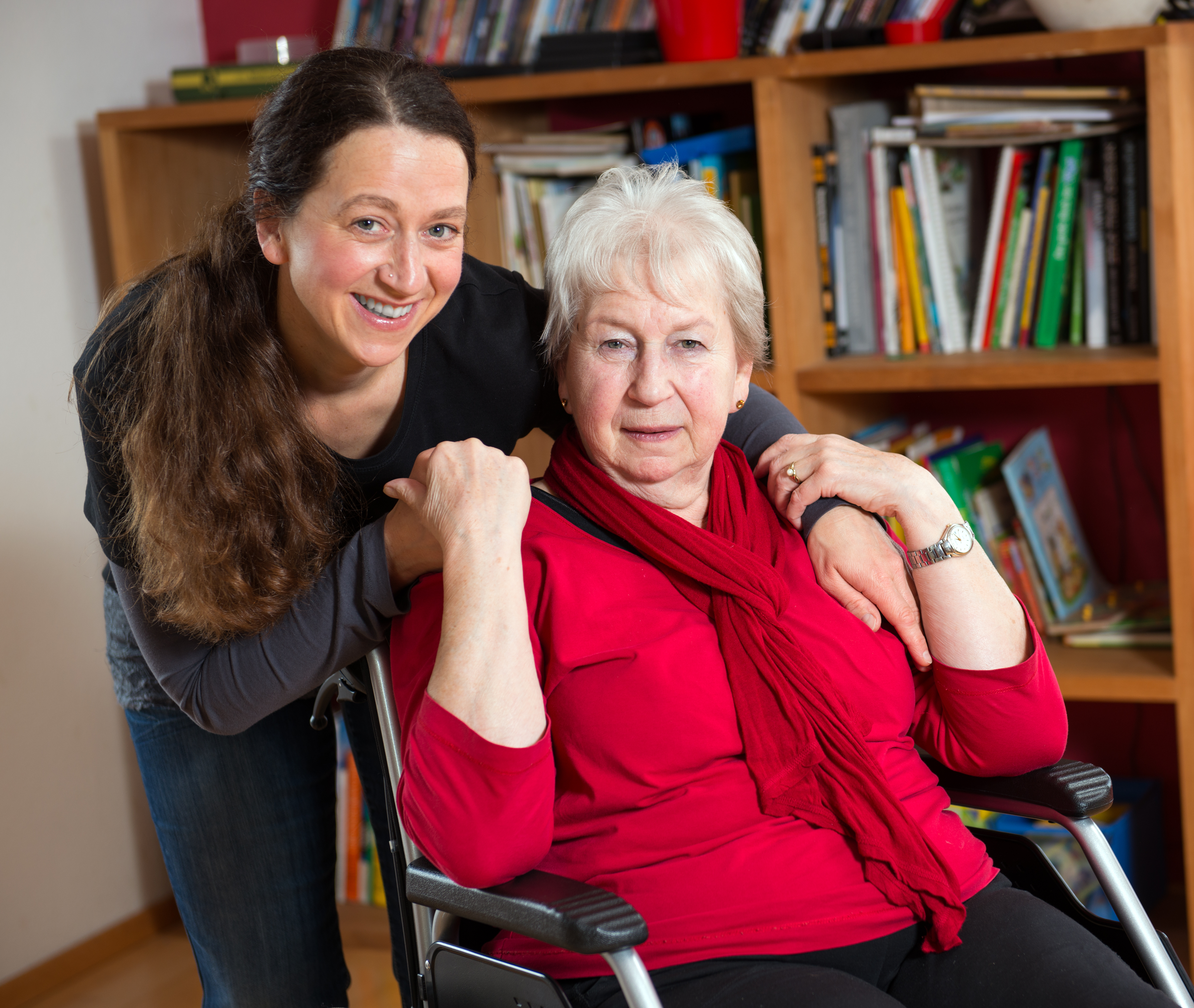 Eine jüngere Frau steht hinter einer älteren Frau im Rollstuhl und legt den Arm um ihre Schulter.