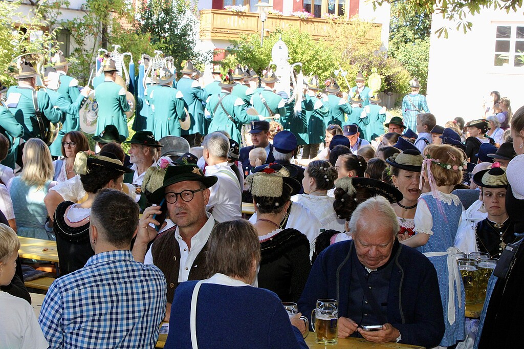 Glockenweihe Beuerberg – Festgemeinde Zahlreiche Festgäste sitzen an Biertischen im Klosterhof. Im Hintergrund steht eine Blaskapelle in einheitlicher Tracht.