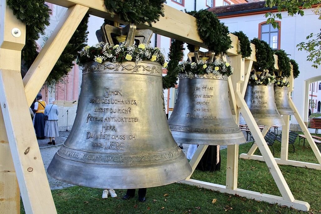 Glockenweihe Beuerberg – Vier neue Bronzeglocken Vier mit grünen Kränzen geschmückte Bronzeglocken hängen nebeneinander in hölzernen Glockenstühlen im Klosterhof Beuerberg.