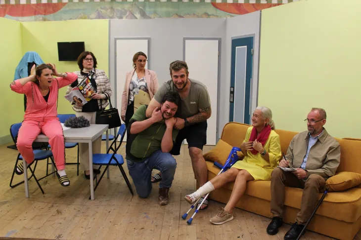 Loisachtaler Bauernbühne – Kulturprogramm Wolfratshausen 2027 Sieben Darsteller der Loisachtaler Bauernbühne in einer lebhaften Theaterszene mit Tisch, Stühlen und Sofa.