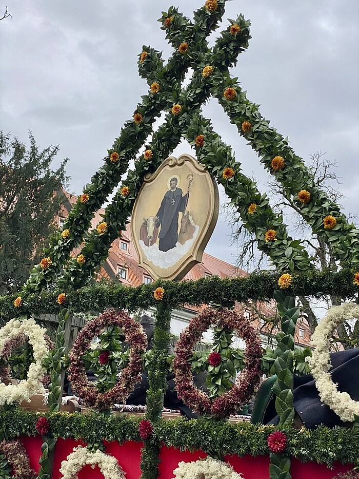 Mit grünen Zweigen und Blumen geschmückter Festwagenaufbau mit einem Bild des heiligen Leonhard, umgeben von Pferdedarstellungen.