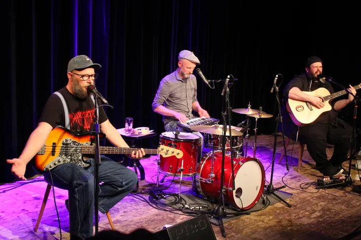 D’Bavaresi – Kulturprogramm Wolfratshausen 2027 Drei Musiker des Musikkabarett-Trios D’Bavaresi sitzen auf einer Bühne mit Bassgitarre, Keyboard, Schlagzeug und Akustikgitarre.