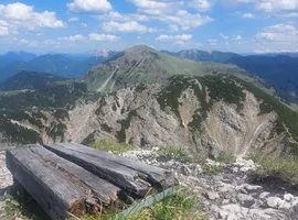 Holzbalken und ein weißer Grenzstein auf einem felsigen Bergkamm, dahinter die Gipfel des Vorkarwendels.