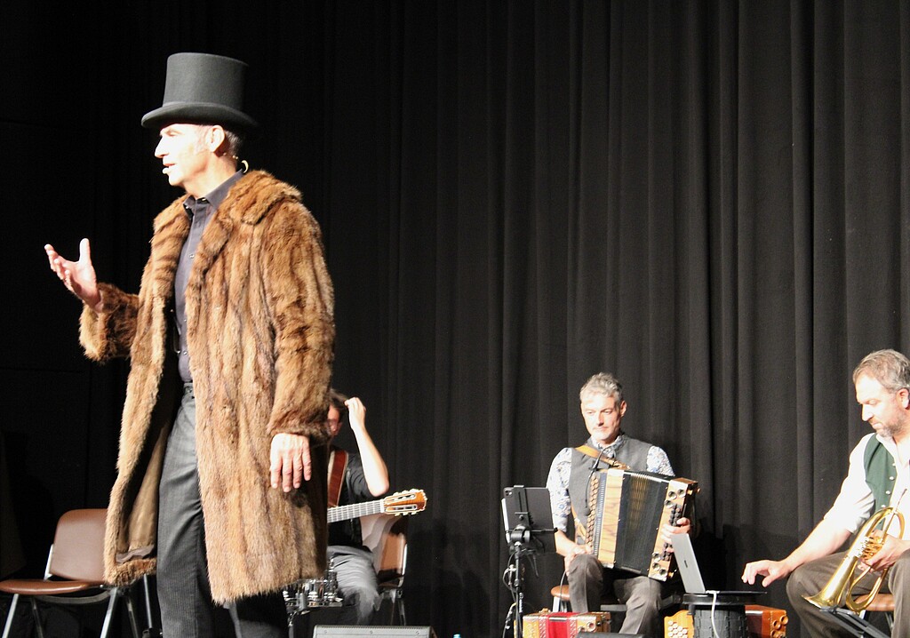 Stefan Kröll bei „Helden und Legenden“ in Wolfratshausen Stefan Kröll steht mit braunem Pelzmantel und schwarzem Zylinder auf der Bühne. Im Hintergrund sitzen Musiker der Kapelle So & So.