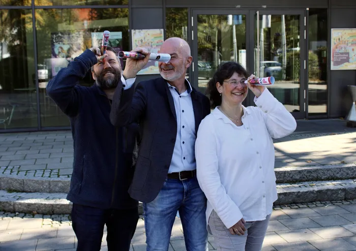 Andreas Kutter, Klaus Heilinglechner und Katharina Roßkopf (von links) blicken lächelnd durch zusammengerollte Programmflyer.