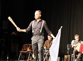 Stefan Kröll steht mit einer Keule in der einen und einer weiß-blau gemusterten Fahne in der anderen Hand auf der Bühne. Im Hintergrund spielen Musiker der Kapelle So & So.