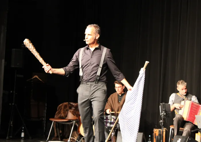 Stefan Kröll steht mit einer Keule in der einen und einer weiß-blau gemusterten Fahne in der anderen Hand auf der Bühne. Im Hintergrund spielen Musiker der Kapelle So & So.