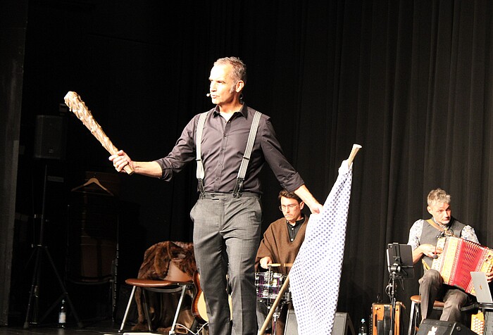 Stefan Kröll mit der Kapelle So & So in Wolfratshausen Stefan Kröll steht mit einer Keule in der einen und einer weiß-blau gemusterten Fahne in der anderen Hand auf der Bühne. Im Hintergrund spielen Musiker der Kapelle So & So.