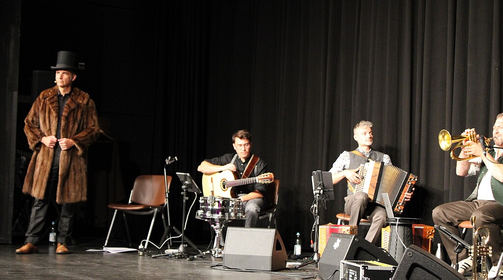 Stefan Kröll und die Kapelle So & So in der Loisachhalle Stefan Kröll steht mit Pelzmantel und Zylinder links auf der Bühne. Rechts sitzen Musiker der Kapelle So & So mit Gitarre, Akkordeon und Blasinstrumenten.