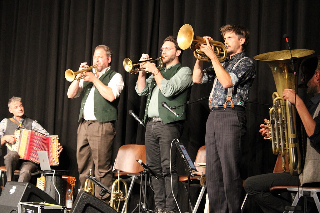 Kapelle So & So bei „Helden und Legenden“ in Wolfratshausen Drei Musiker der Kapelle So & So spielen stehend auf ihren Blechblasinstrumenten. Links sitzt ein Akkordeonspieler, rechts ist ein weiterer Musiker mit einem großen Blasinstrument zu sehen.