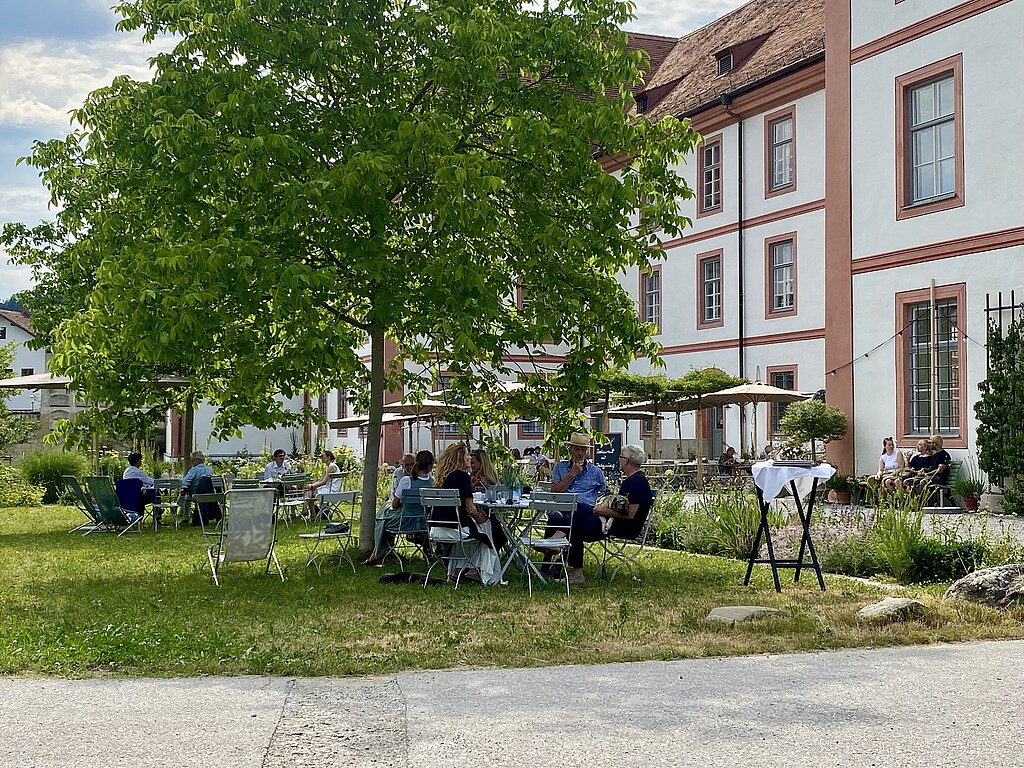 Klostergarten Beuerberg Besucherinnen und Besucher sitzen im Garten des Klosters Beuerberg an Tischen unter einem Baum.