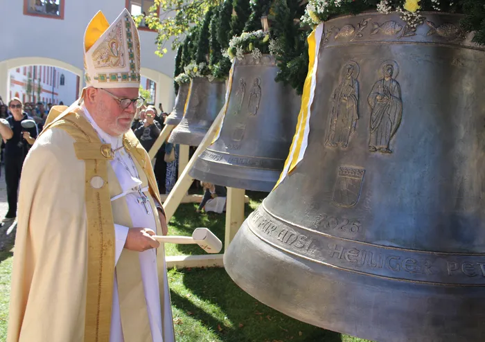 Kardinal Reinhard Marx steht mit einem Schlägel vor einer der vier neuen Bronzeglocken der Stiftskirche St. Peter und Paul in Beuerberg.