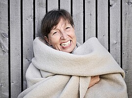 Porträt der Couturière Constanze Janusch in einem hellen, voluminösen Mantel vor einer Holzwand